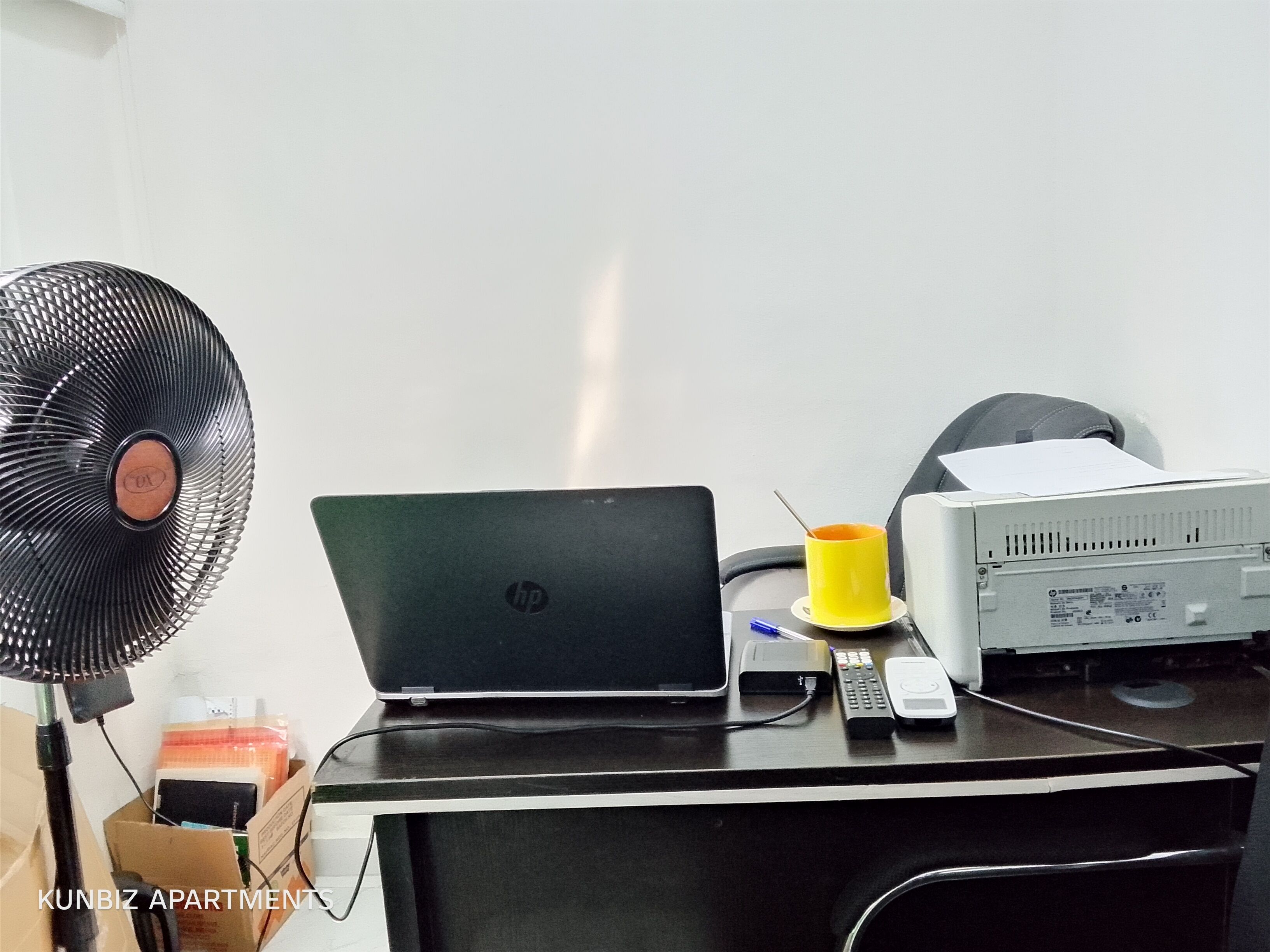 Un bureau avec un ordinateur portable, un ventilateur, une imprimante et une tasse.