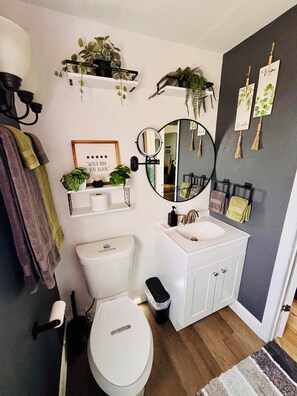 Ein modernes Badezimmer mit Toilette, Waschbecken und Spiegel. Pflanzen stehen auf Regalen und ein Handtuchhalter mit Handtüchern ist vorhanden.