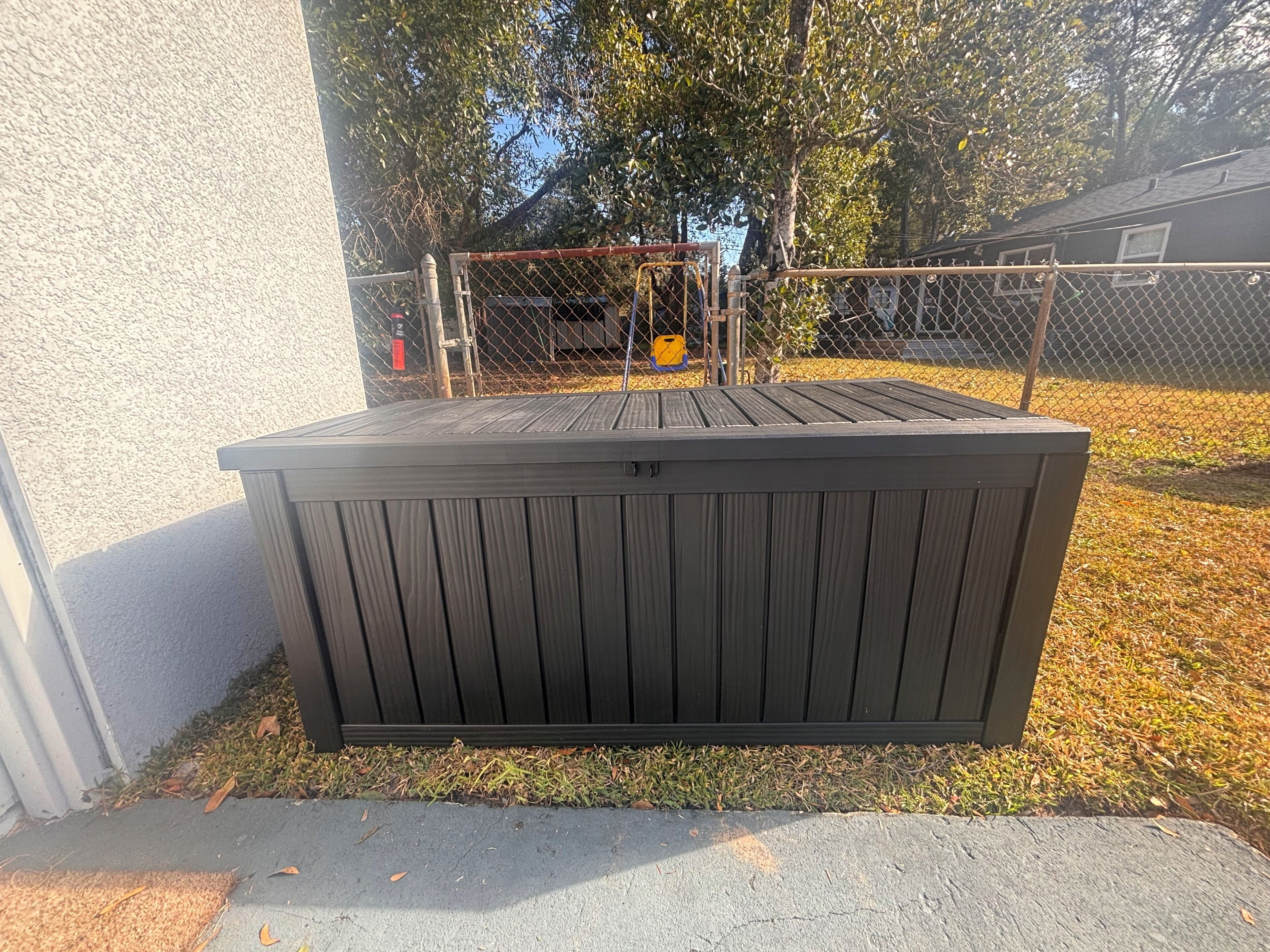 Un abri de jardin noir, situé à l’extérieur, avec une clôture en métal, adossé à un mur blanc et donnant sur une zone herbeuse.