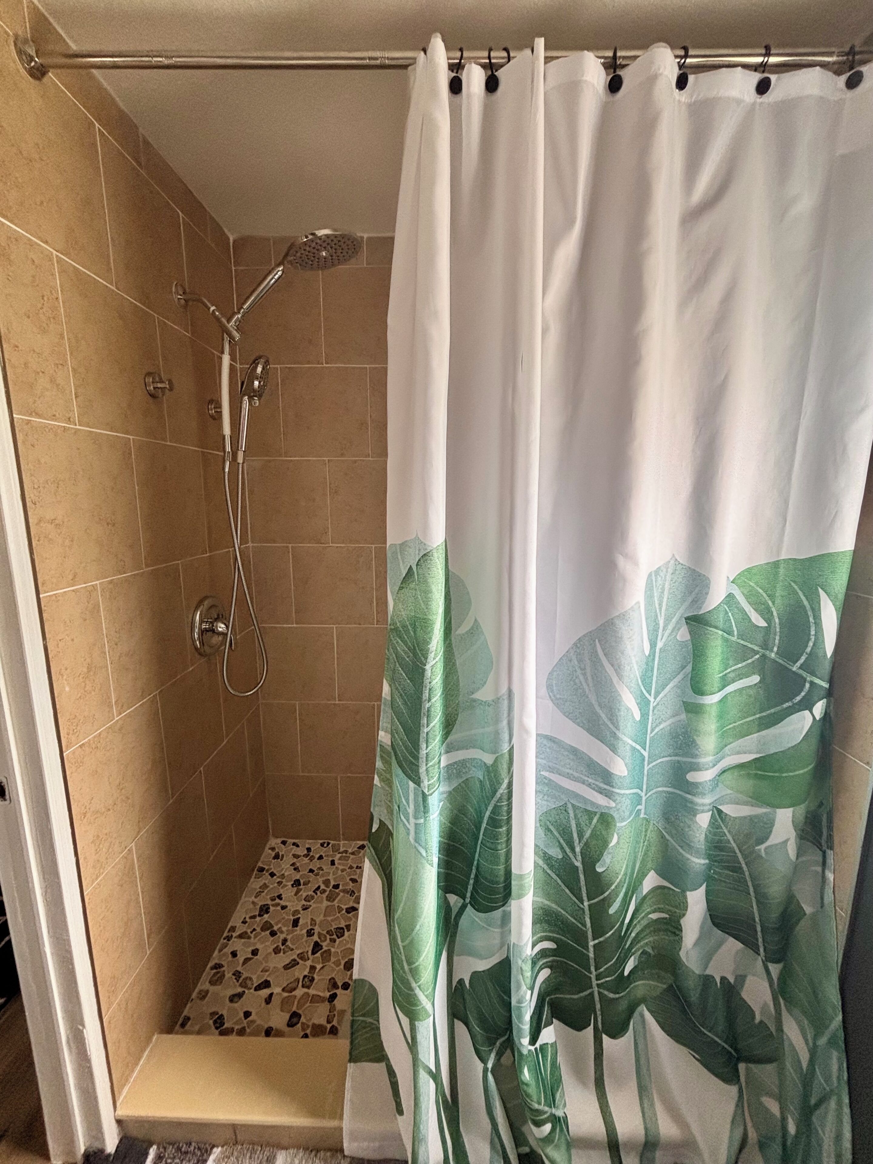 A shower with a white curtain featuring a green leaf pattern, a showerhead, and a tiled wall.