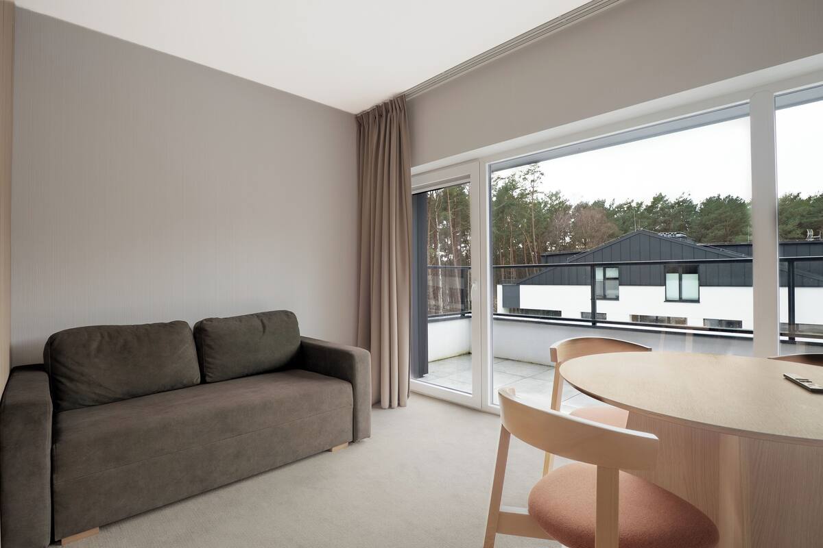 A modern living room with a grey sofa, a round wooden table, and a large sliding glass door leading to a balcony.