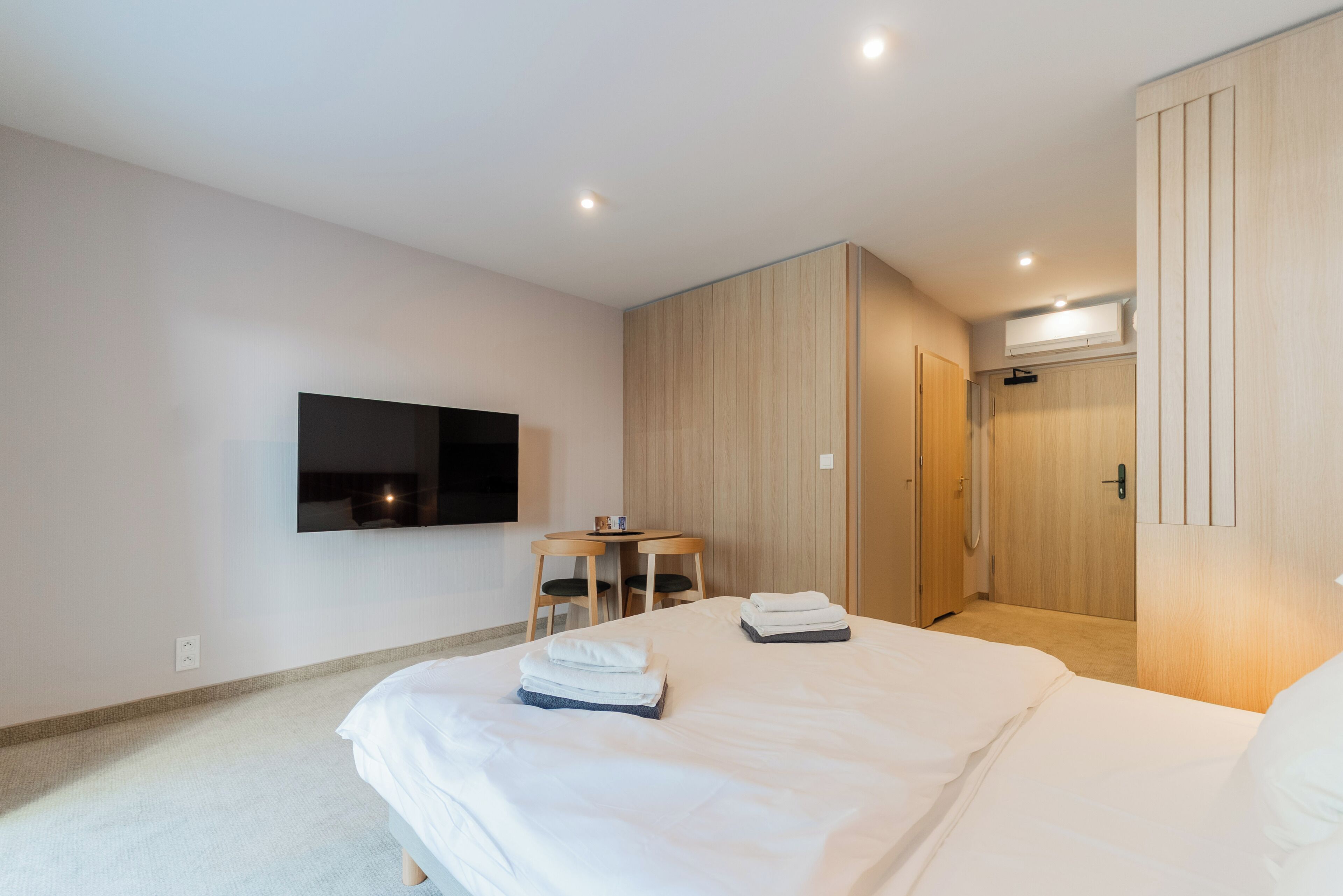 A modern hotel room with a large bed, a flat-screen TV mounted on the wall, a small round table with chairs, and a wardrobe with sliding doors.