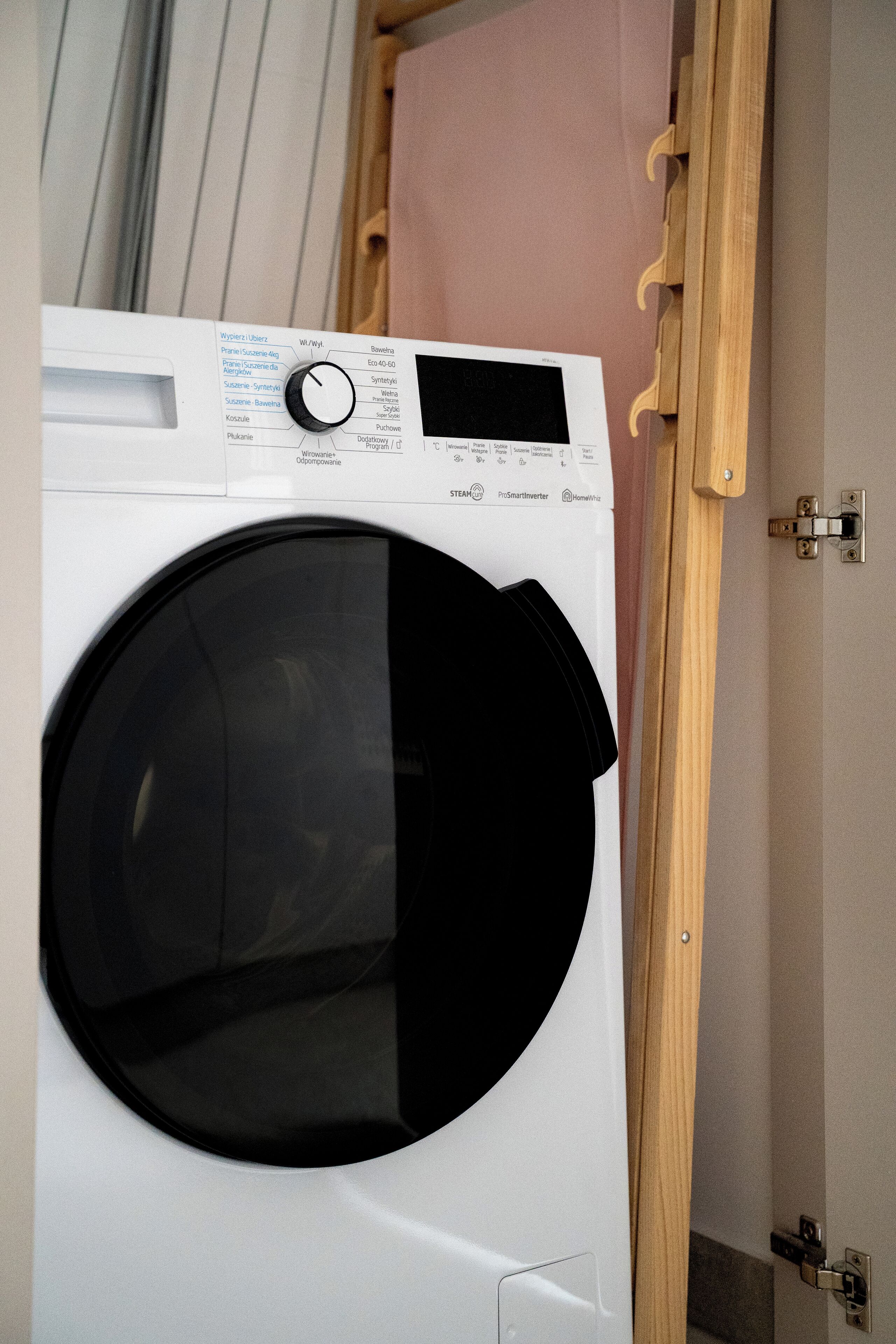 A front-loading washing machine with a digital display and control panel, located in a room with wooden doors and a light-colored wall.
