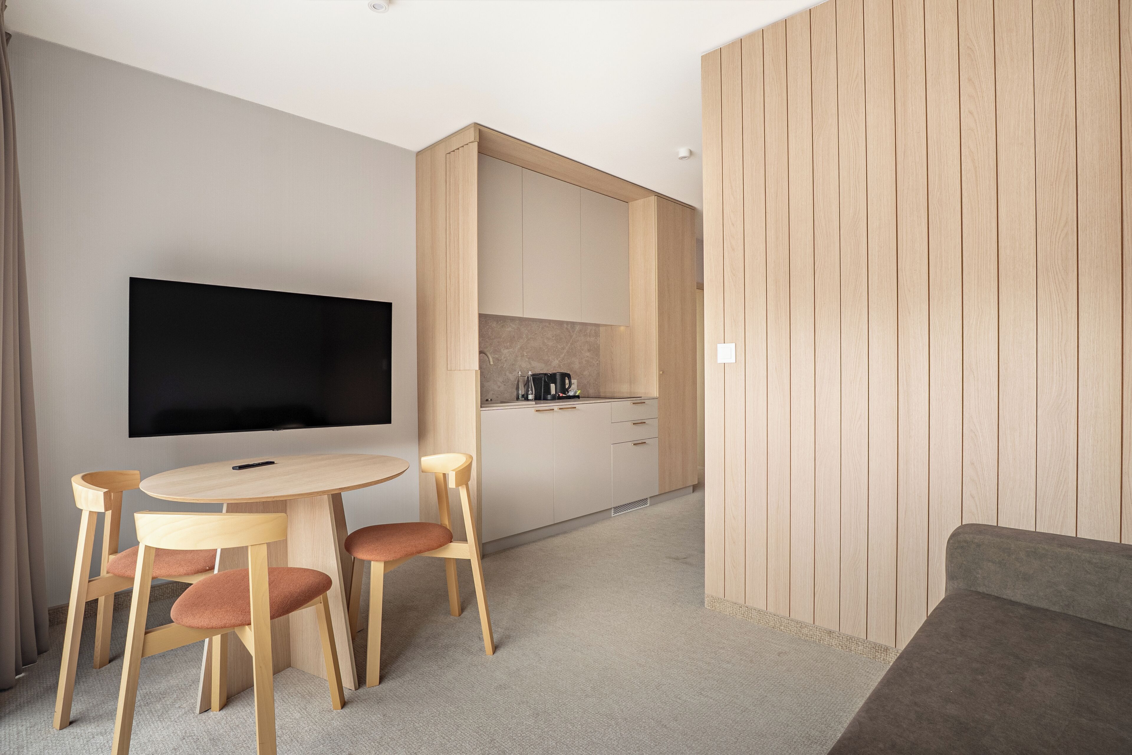 A modern living room with a flat-screen TV, wooden furniture, and a kitchen area in the background.