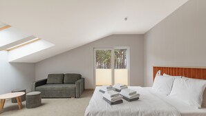 Une chambre moderne avec un grand lit, un canapé, une petite table et une fenêtre donnant sur des arbres.