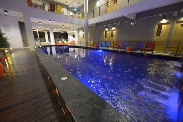 An indoor swimming pool with a wooden deck area and lounge chairs. - Brick House Hostel Chiangmai (Chiang Mai)