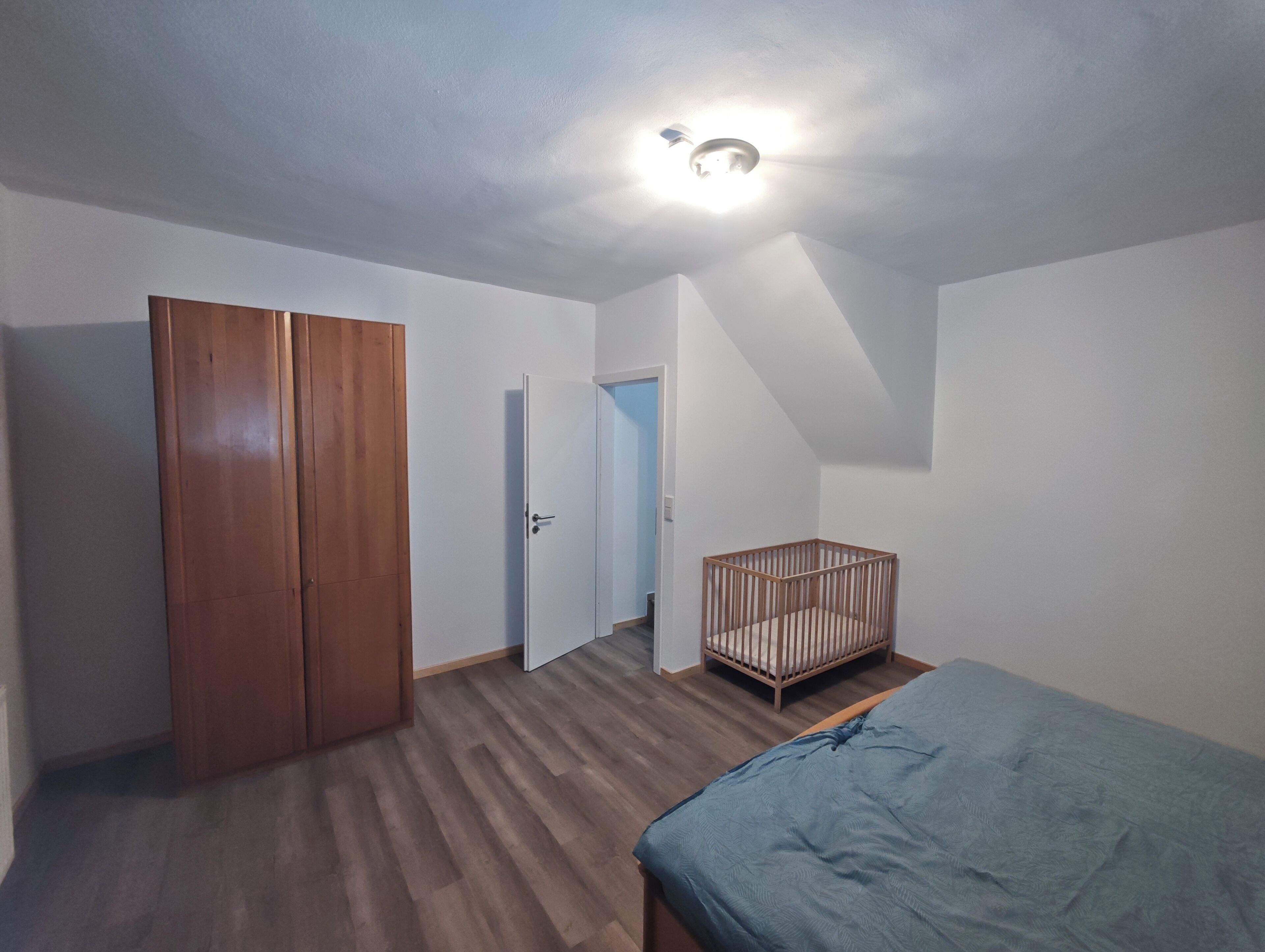 A room with a wooden wardrobe, a crib, and a bed.