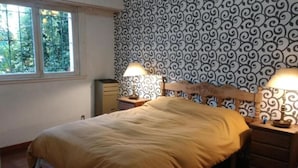 A bedroom with a bed, two bedside tables with lamps, a dresser, a window with a view of greenery, and patterned wallpaper.