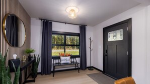 Una habitación moderna con una puerta negra, un banco con un cojín que dice 'Be Our Guest', un perchero y vistas al verdor exterior.