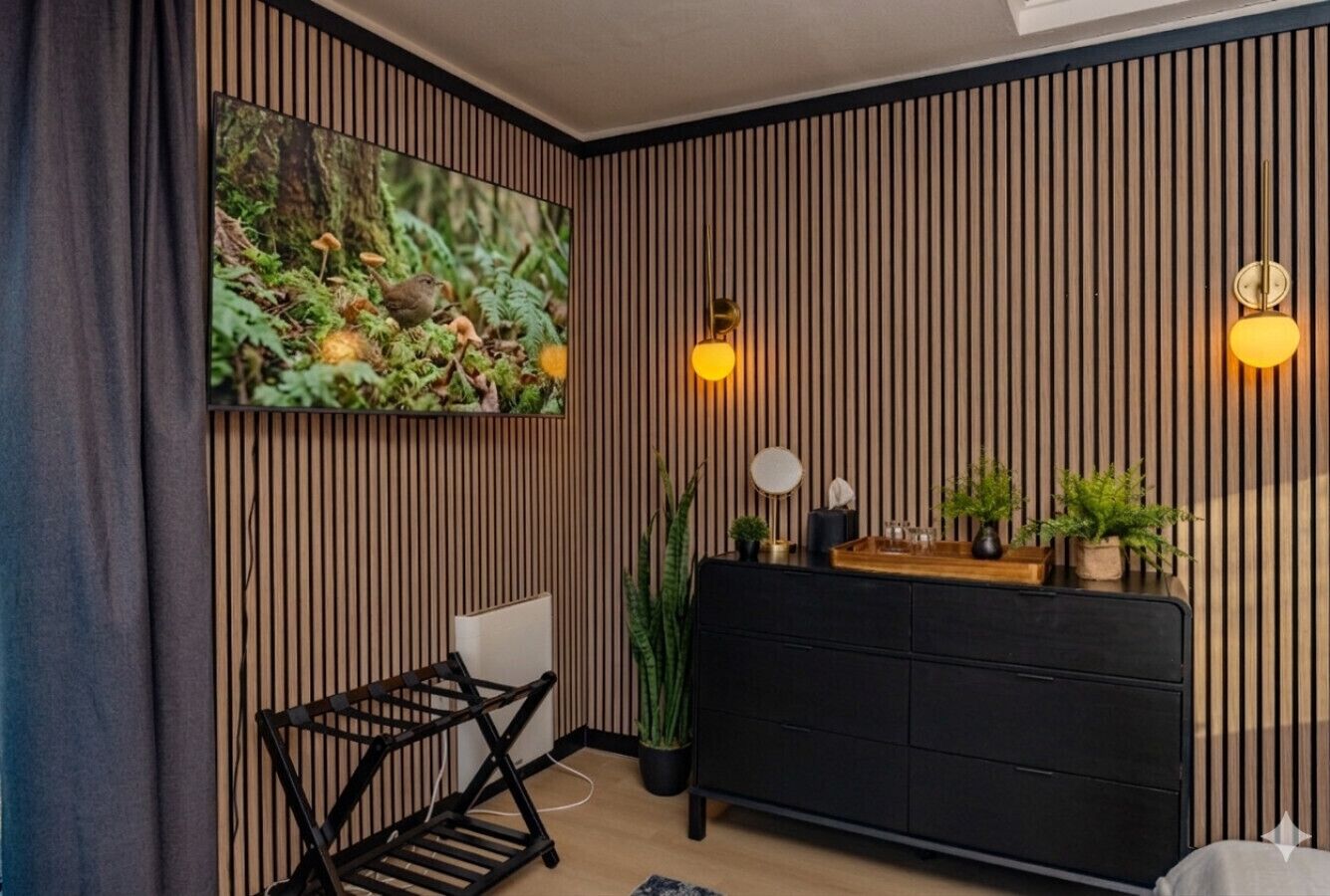 Ein modernes Zimmer mit einer schwarzen Couch, einer Wand aus Holzlamellen, einem großen gerahmten Bild und zwei Wandleuchten.