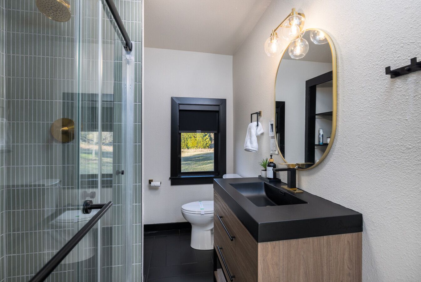A modern bathroom with a glass shower enclosure, a black sink with a wooden vanity, a large mirror with a gold frame, and a view of greenery outside.