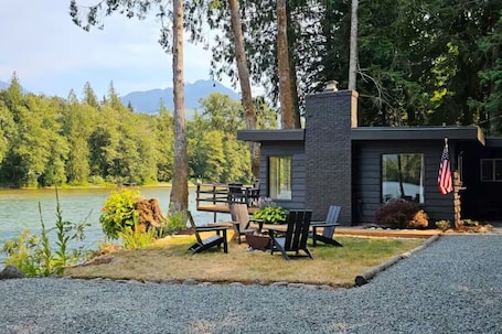 A modern house with a deck overlooking a lake, surrounded by trees and mountains.