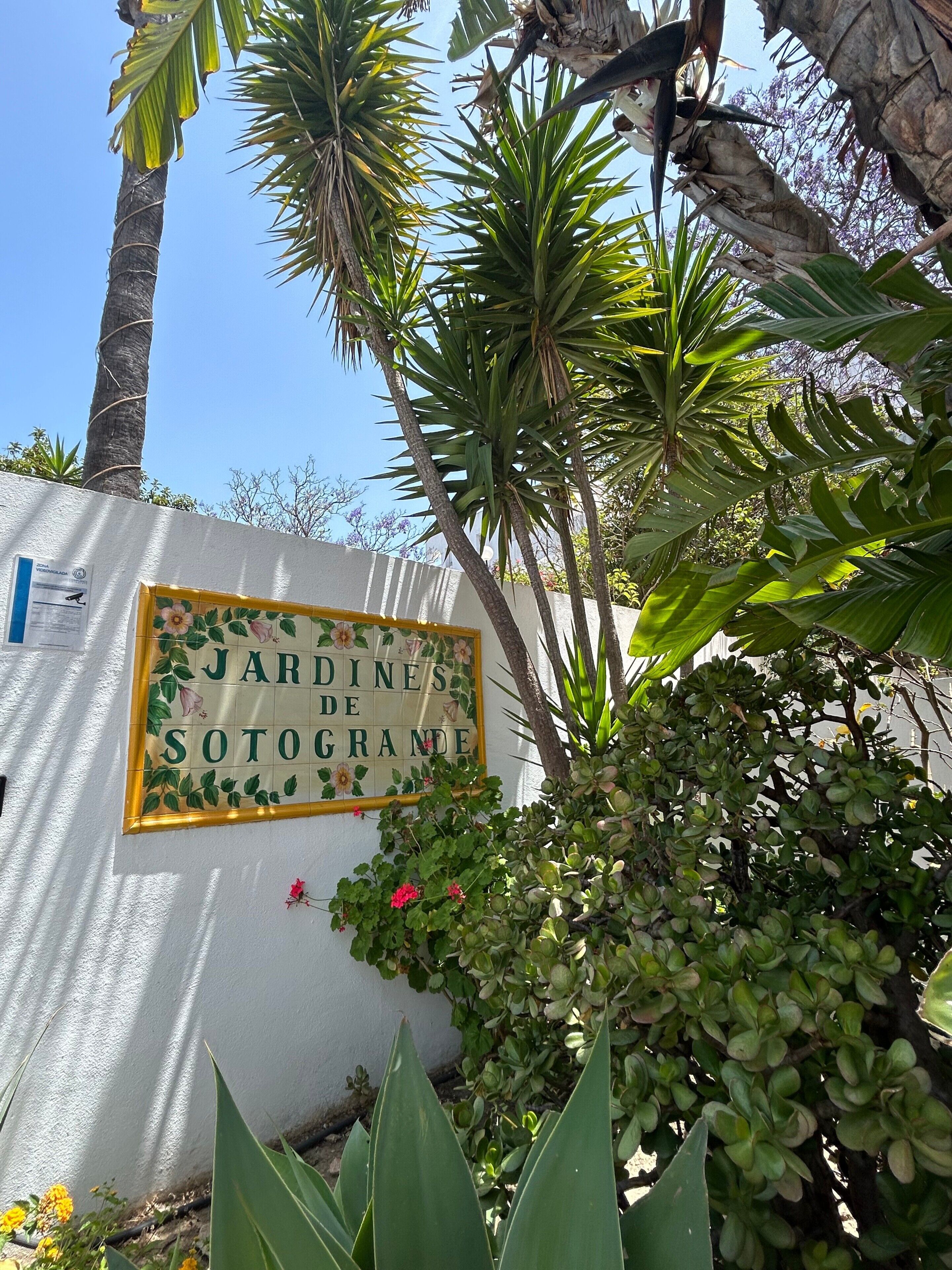Une enseigne portant l’inscription « JARDINES DE SOTOGRANDE », entourée de plantes tropicales et adossée à un mur blanc.