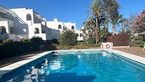 A swimming pool with clear blue water, surrounded by white buildings with arched windows and a palm tree. - Duplex 'Sotogrande' with Shared Pool, Terrace and Wi-Fi (Sotogrande)