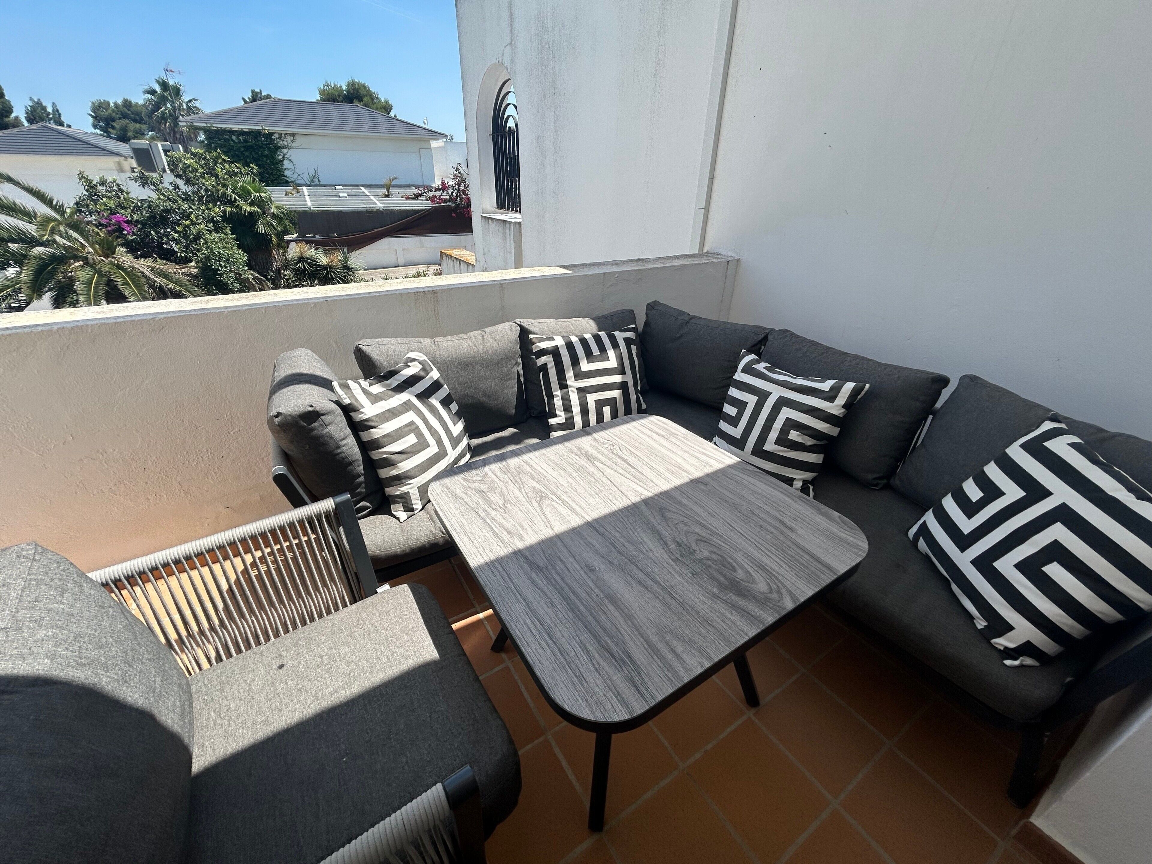 Un balcone con un divano grigio, un tavolo di legno e un cuscino decorato.