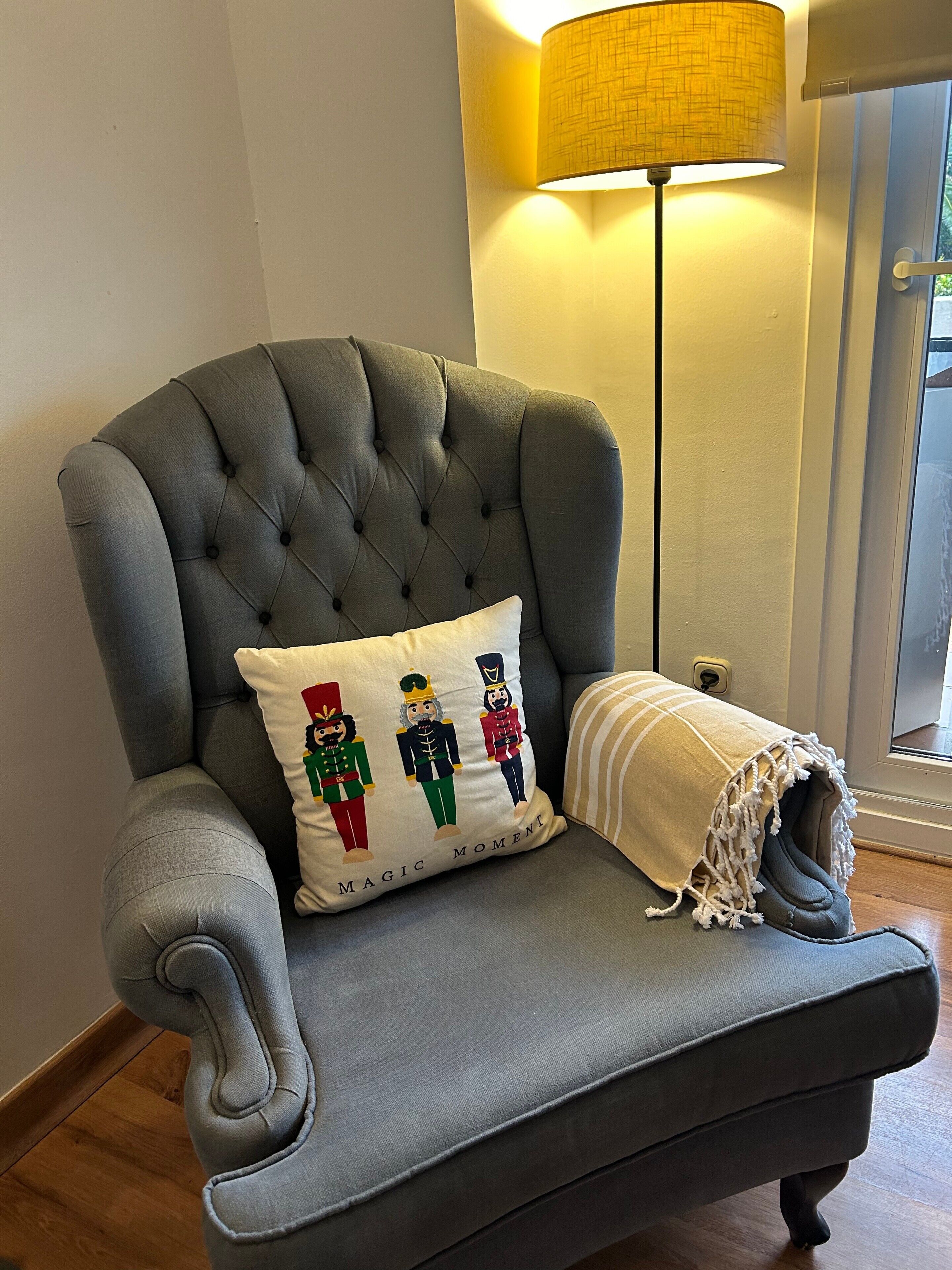 A grey tufted armchair with a decorative pillow featuring nutcracker figures, positioned near a lamp and a window.