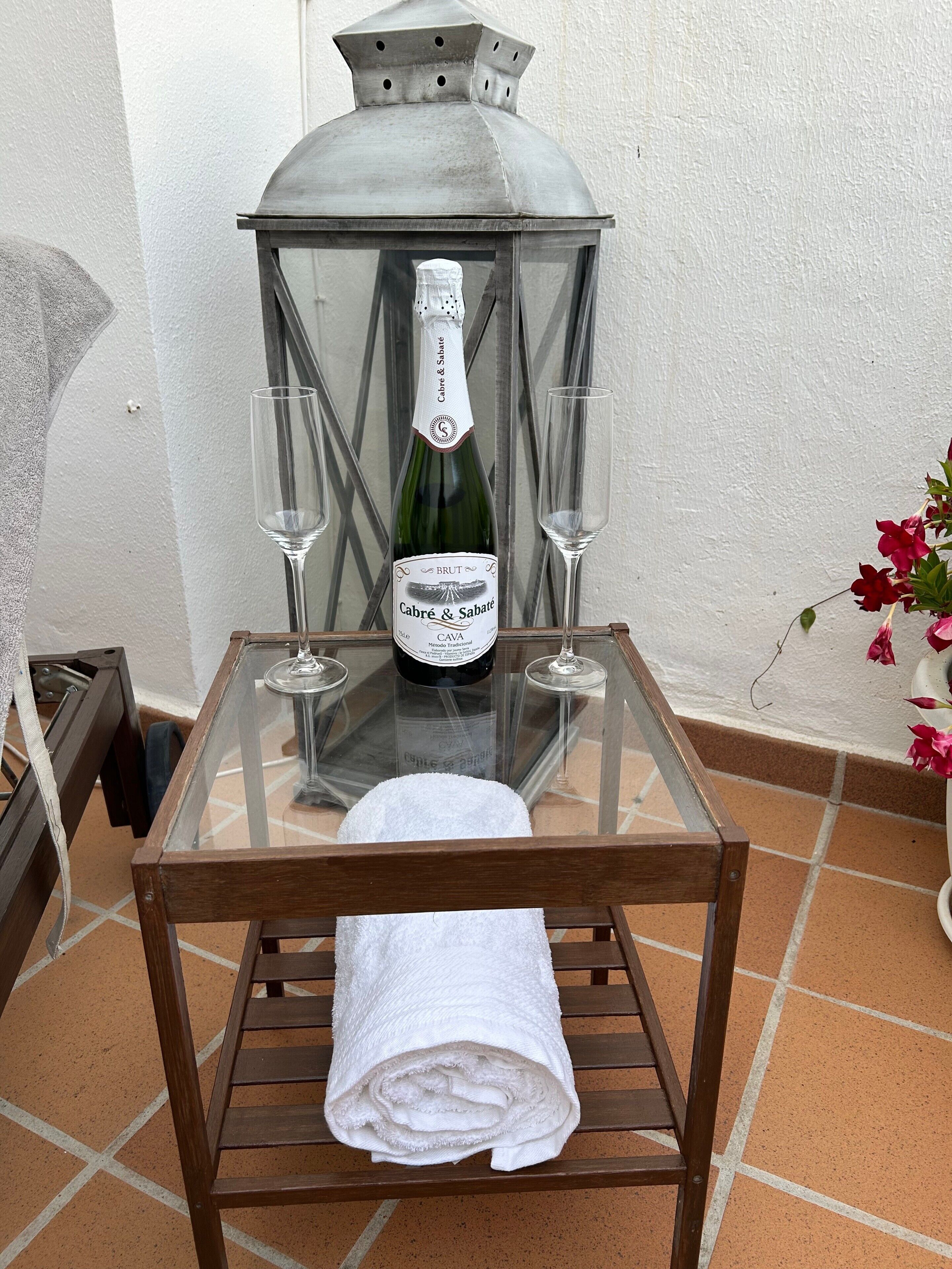 Une table en verre avec une bouteille de CAVA Cabre & Sabate, deux verres et une serviette blanche roulée.