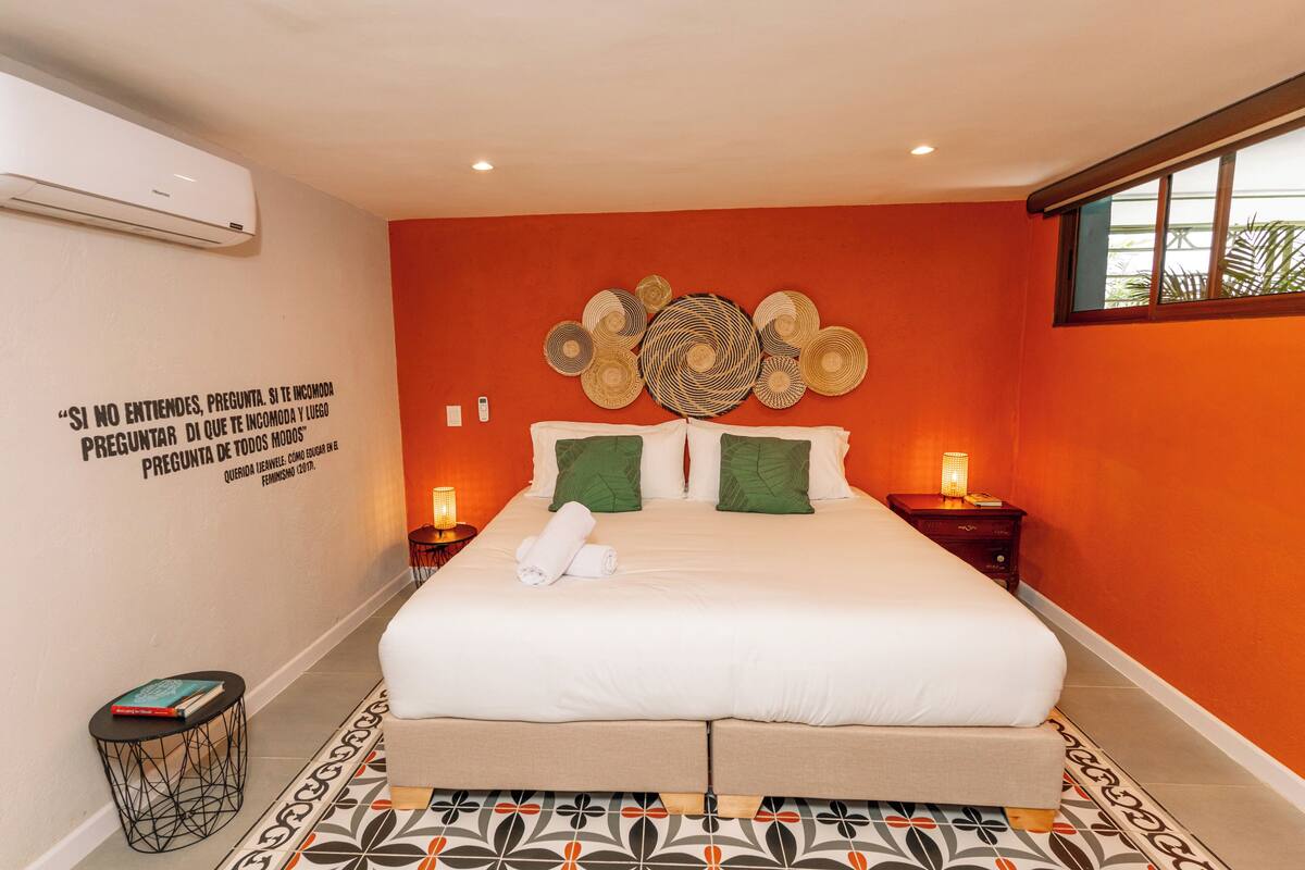 A hotel room with a large bed, green pillows, a decorative wall art piece, and a small bedside table with a lamp.