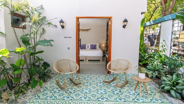 Ein Zimmer mit einem Bett, zwei Korbstühlen, einem kleinen Tisch mit Blumentopf und einem Fliesenboden mit geometrischem Muster.