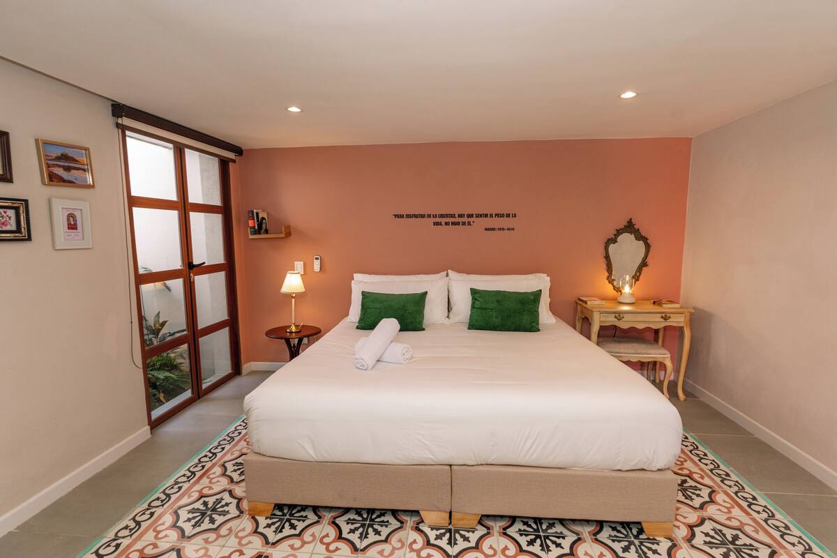 A bedroom with a large bed, a bedside table with a lamp, a mirror, and a wall quote.