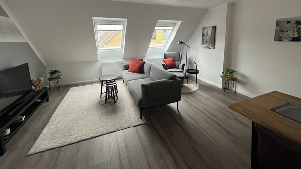 Un moderno apartamento tipo loft con un sofá gris, una pequeña mesa redonda, una lámpara de pie negra y una mesa de centro de madera. Hay dos ventanas con tragaluces, un radiador y un cuadro en la pared.