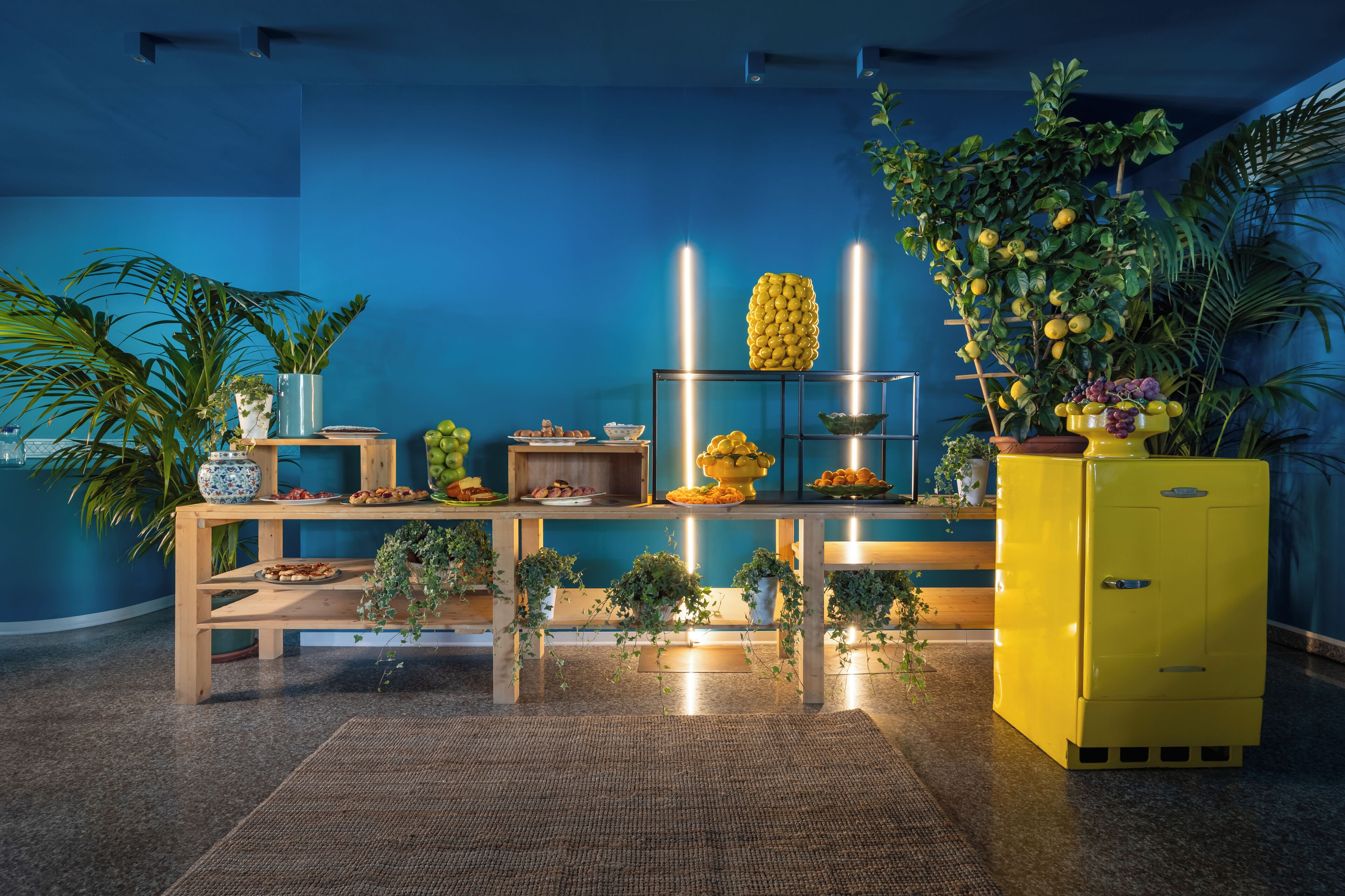 A room with a blue wall, a wooden table with food, a yellow cabinet, and potted plants.