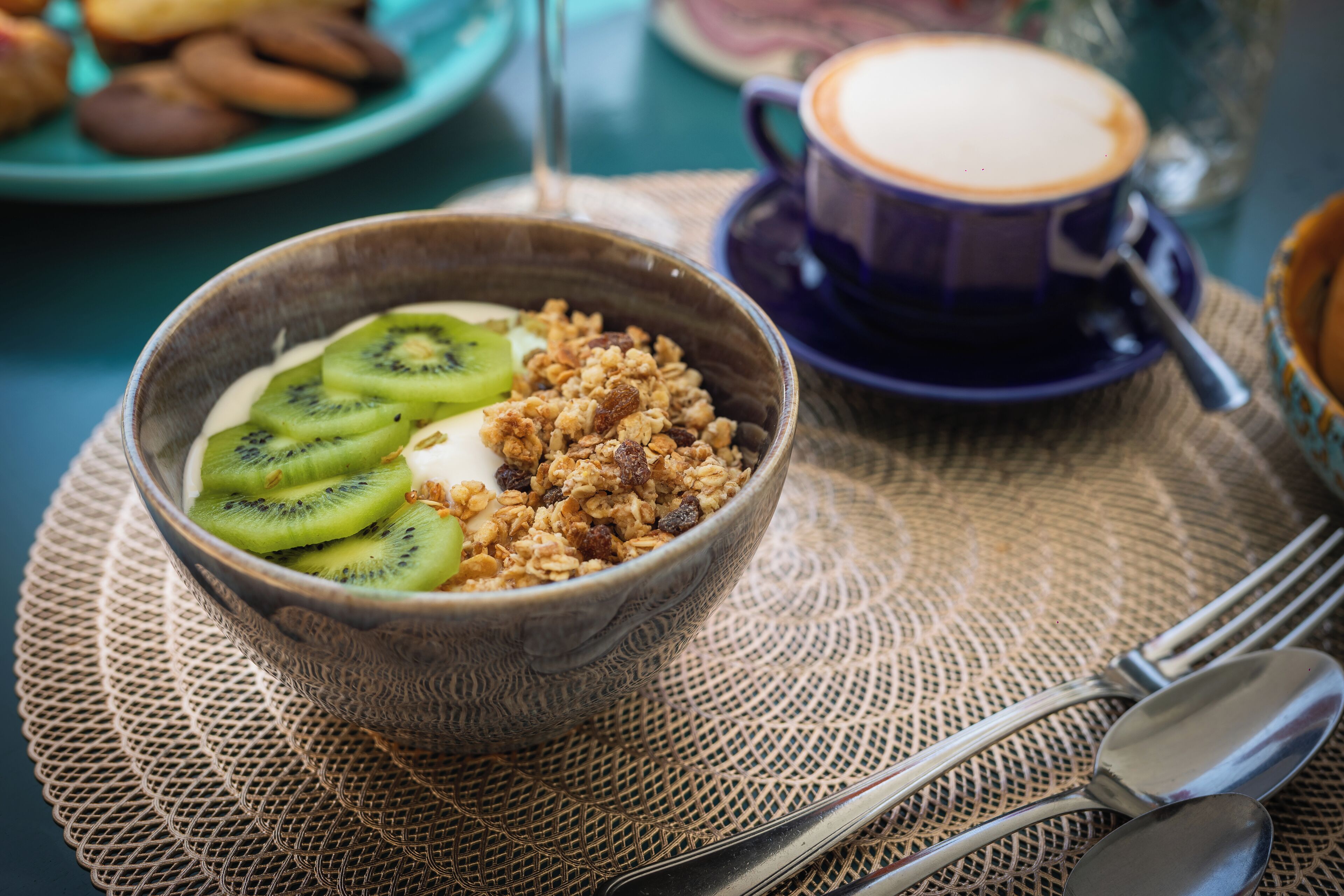 Un bol de céréales garni de tranches de kiwi et de granola, posé sur un set de table texturé, accompagné d’une tasse de café et d’une cuillère.