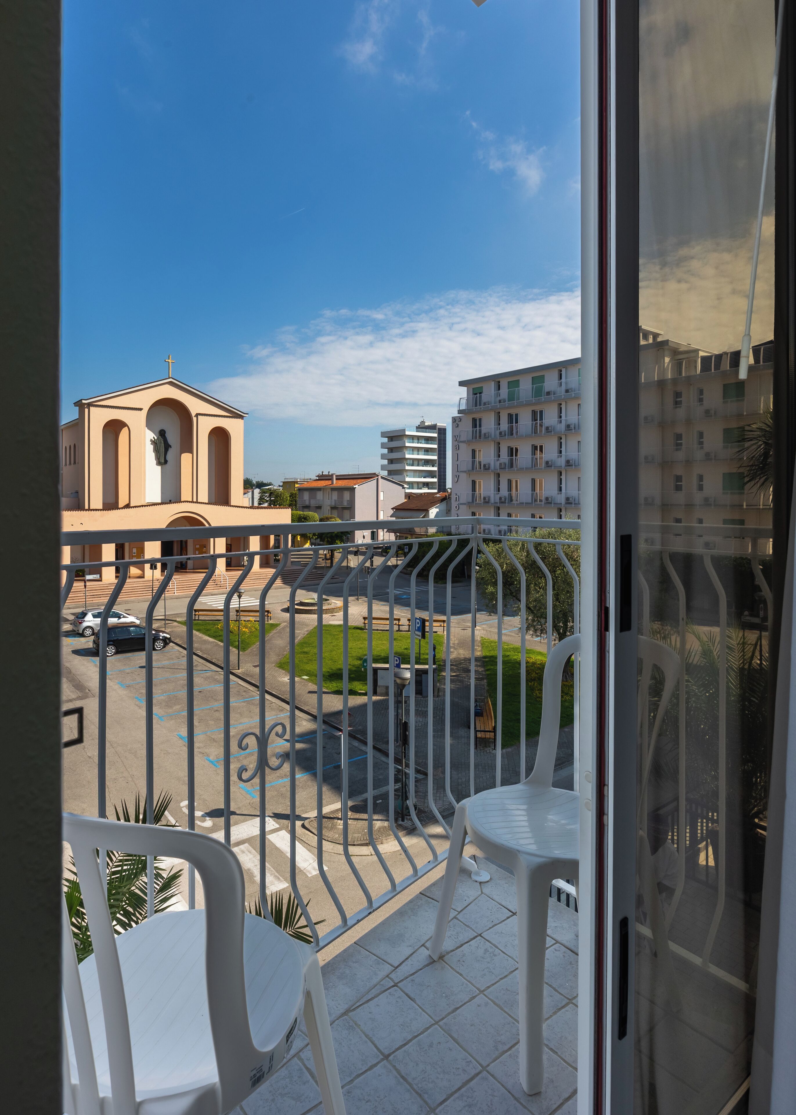A balcony with white chairs, a view of a church, and a parking area.