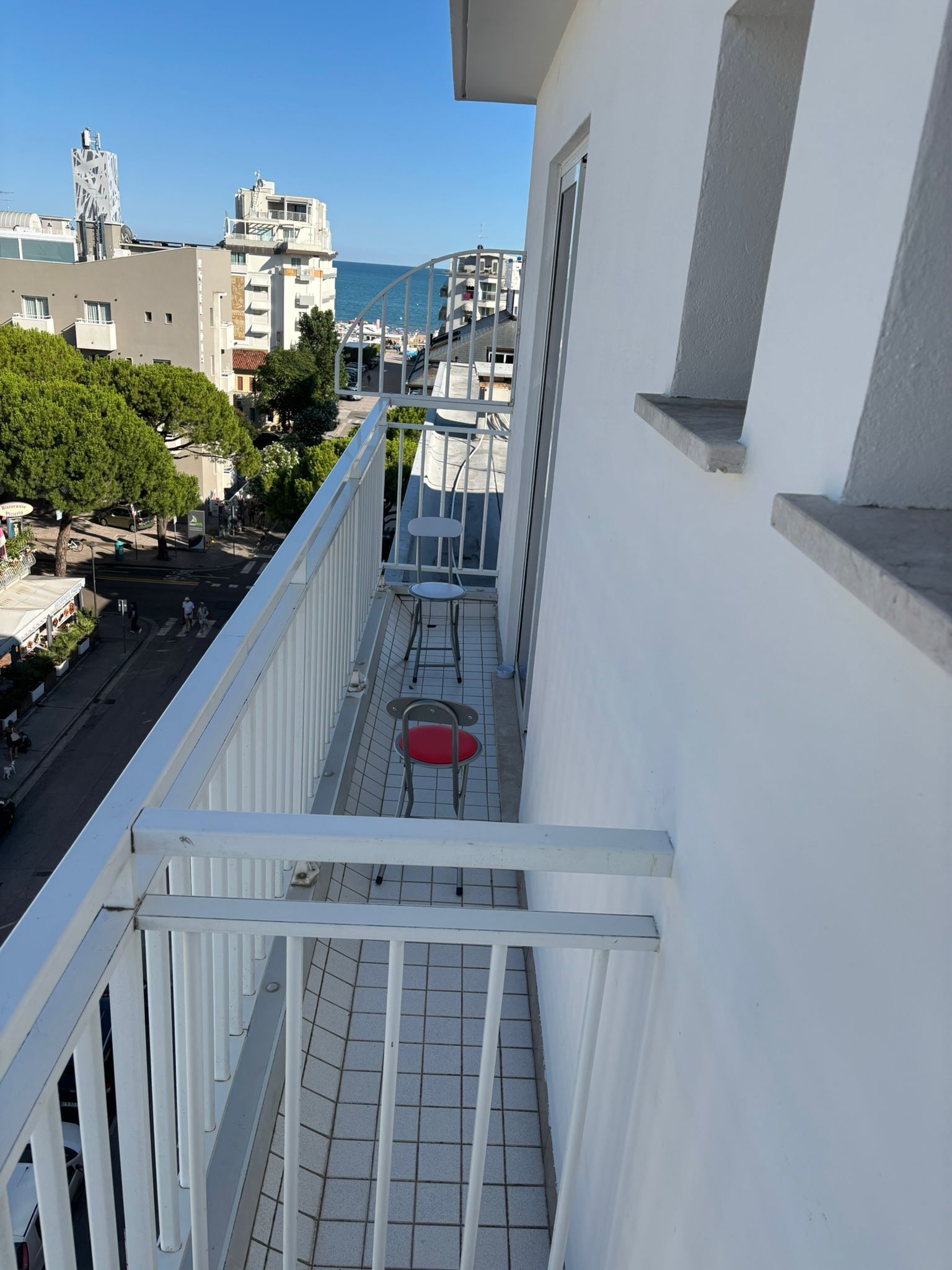 A balcony with a red chair and a small table, overlooking a street with buildings and trees.
