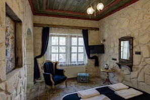 A room with a stone wall, a bed with white linens, a blue armchair, a small table with a kettle, and a mounted television. - cappadocia keyfiala hotel (Urgup)