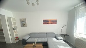A modern living room with a grey sectional sofa, a wooden coffee table, a bookshelf, a painting, and a floor lamp. - House with courtyard, 3-minute walk from the beach 🏖️ - the sea breeze 🌊 (Gravelines)