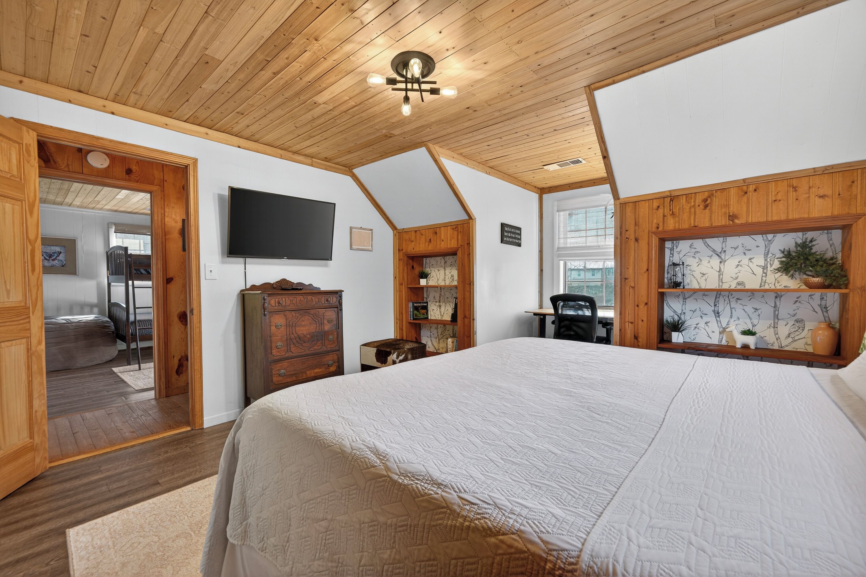Quarto com cama grande, teto de madeira, TV na parede e janela com vista para o exterior.