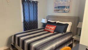 A neatly made bed with striped bedding, a pillow with a red tag, a nightstand with a lamp, and a painting of a coastal scene on the wall.
