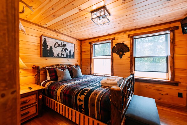Une chambre dans une cabane en bois, avec un lit, une table de chevet, un banc, un ventilateur fixé au mur et une fenêtre donnant sur la forêt.