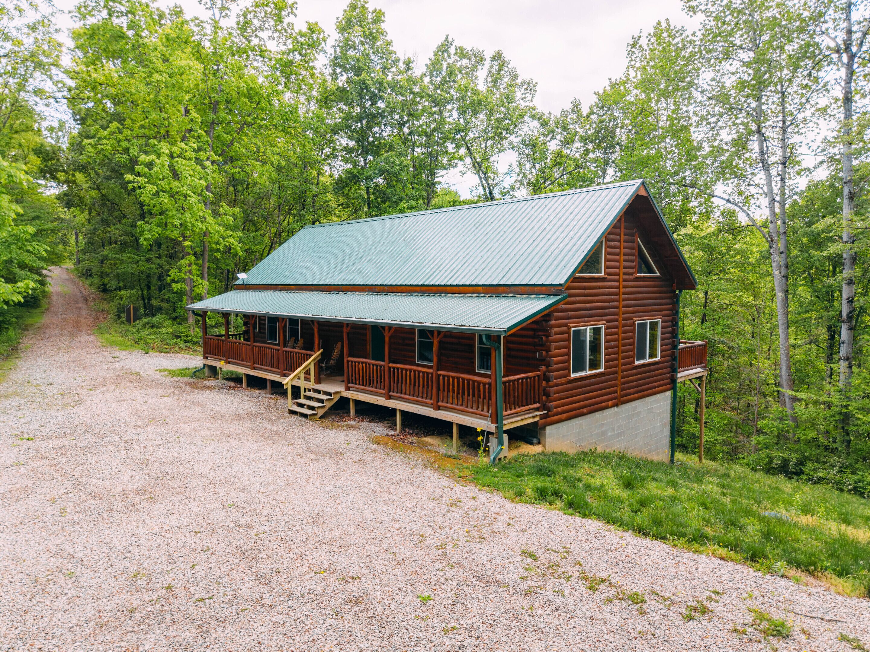 A wooden cabin with a porch, a gravel driveway, and a forested backdrop.