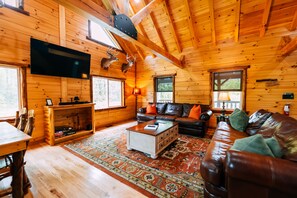 Interior de una cabaña de troncos con techo de madera, sofá de cuero, televisor y chimenea.