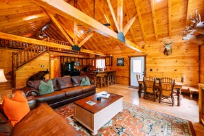 Interior de una cabaña de troncos con escalera de madera, sofá de cuero, mesa de comedor y sillas.
