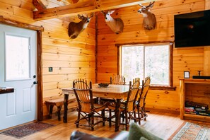 Interior de una cabaña de madera con una mesa de comedor, sillas, un televisor y venados montados en las paredes.
