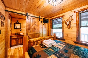 Het interieur van een houten hutje, met een bed, een houten bankje, een opgeplakt dierenhoofd, een televisie en een raam met uitzicht op de omgeving.