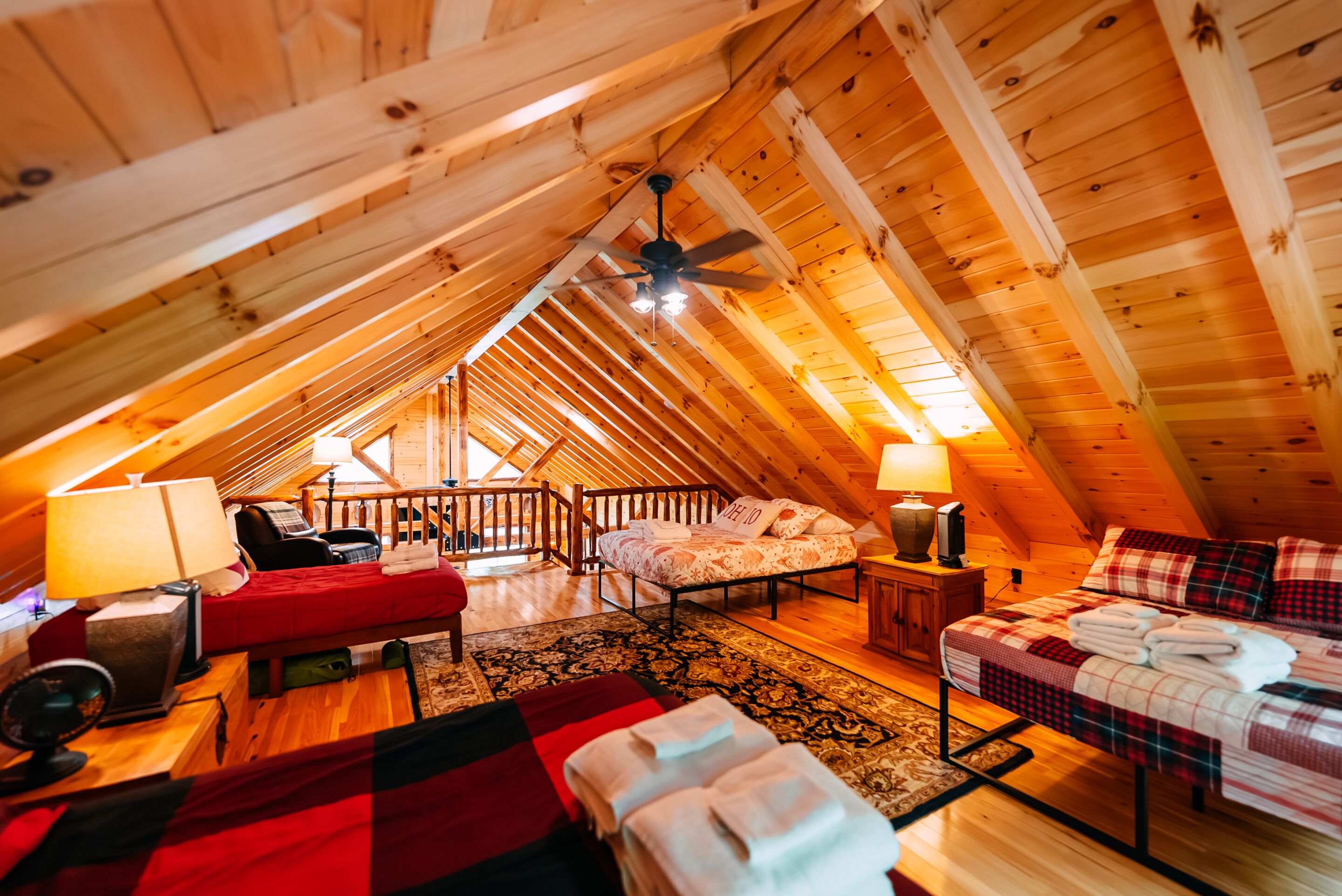 An attic bedroom with a sloped ceiling, a ceiling fan, two beds, a red sofa, a wooden floor, and a patterned rug.
