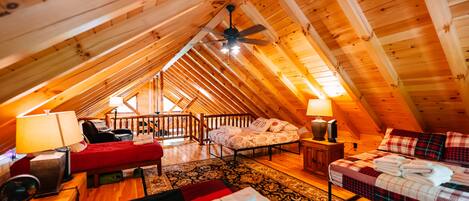 An attic bedroom with a sloped ceiling, a ceiling fan, two beds, a red sofa, a wooden floor, and a patterned rug.