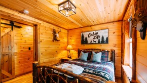 Et interiør af en træhytte med en seng, en lampe, et skilt med teksten 'Cabin Life' og en hjortehoved dekoration.