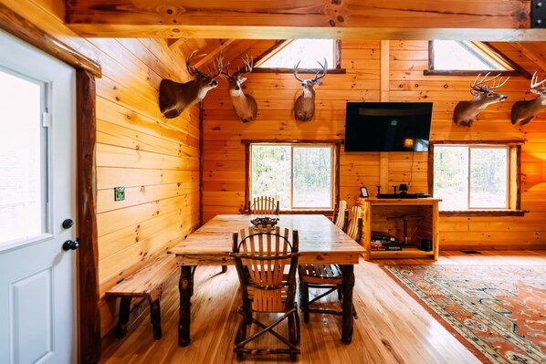 Interior de una cabaña de troncos con una mesa de comedor de madera, sillas, ciervos montados como trofeos, una chimenea y un televisor.