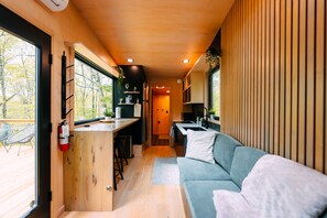 A compact, modern interior with a kitchenette, seating area, and large windows offering a view of the outdoors. - Sangja | Hocking Hills, 60 Acres, Modern, Hot Tub (Logan)