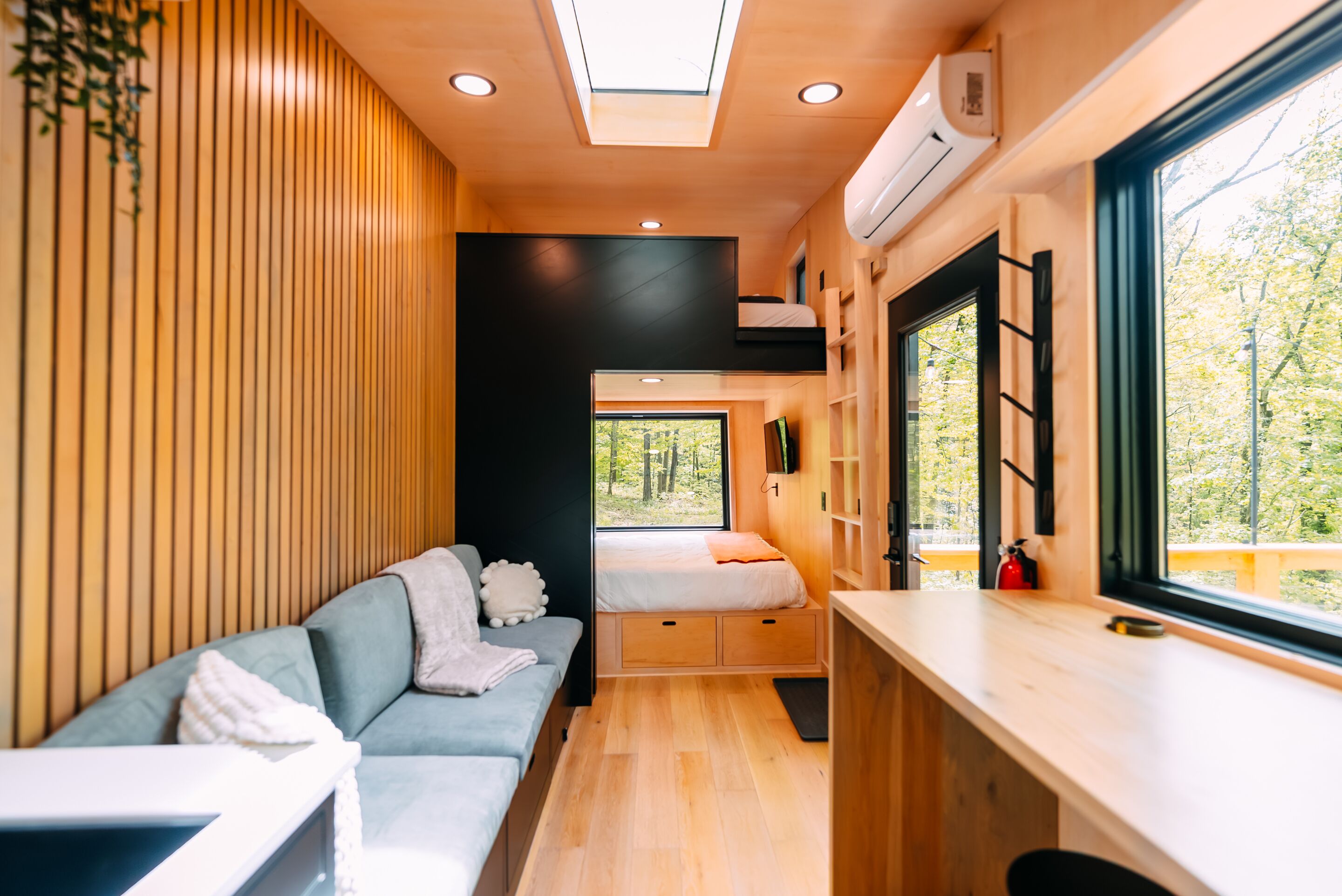 Een compact interieur met een bank, een bed, een kitchenette en een raam met uitzicht op bomen.