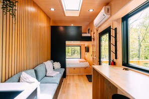 Un intérieur compact comprenant un canapé, un lit, un coin cuisine et une fenêtre donnant sur la forêt.
