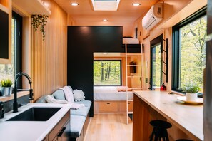 Un intérieur compact et moderne, doté d’un lit superposé, d’une kitchenette et de grandes fenêtres offrant une vue sur les arbres.