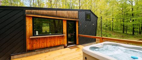 A wooden deck with a hot tub, a wooden cabin with a sliding door, and a forested backdrop.