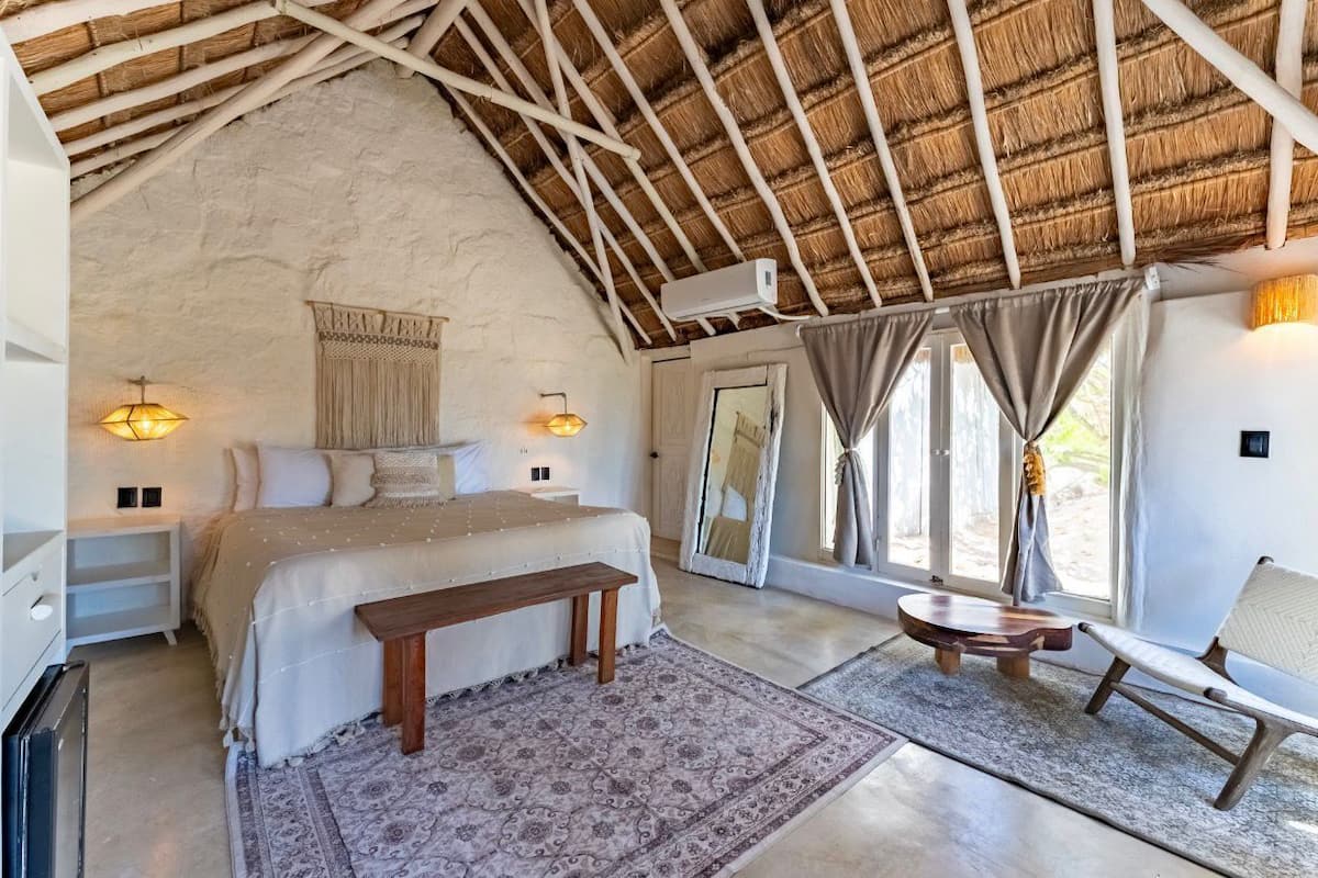 Une chambre à coucher avec un toit de chaume, un lit, des tables de chevet, un banc, une chaise et une grande fenêtre avec des rideaux.