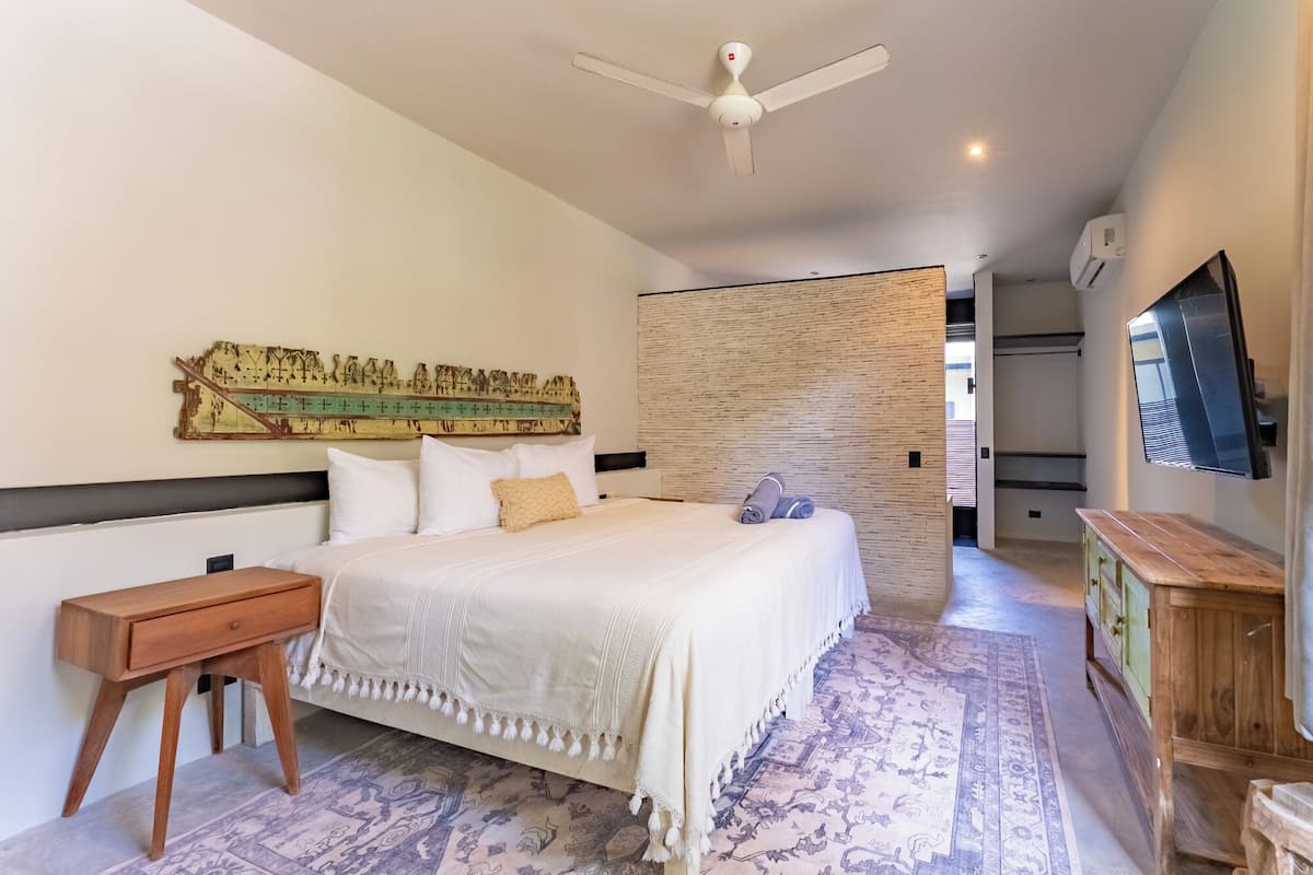 Une chambre d’hôtel avec un lit, une table de chevet en bois, un téléviseur fixé au mur, un ventilateur de plafond et un tapis à motifs.